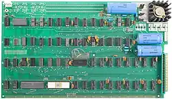 A large, rectangular circuit board with mostly uniform chips arranged neatly in a grid. The rows are labeled A through D and the columns are numbered 1 to 18. Printed between rows of chips is the text, "Apple Computer 1", "Palo Alto, [California] Copyright 1976". There are three large cylindrical capacitors laying sideways in the corner. The board is sprinkled with small components including ceramic resistors and jumper wires.