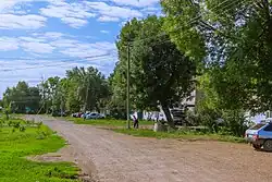 A photograph of a street within Tugay. The road is made of dirt. Houses on the right-hand side of the street are obscured by tall trees.