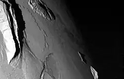 A portion of a planetary body with a pair of large, mountainous ridges on the left side of the image, a shorter, rugged domical mountain at top center, an elliptical pit near bottom center, and the boundary between the dayside (to the left) and the nightside (to the right) running down the right side of the image. Two small mountain peaks are seen near this boundary at lower right.