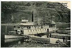 A steamboat waits at a pier on a river bank while sacks of cargo are loaded.
