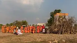 Ritual procession of Buddhist monks in front of a coffin over a pyre before lighting the fire, during funerals in the countryside of Don Det. Holding a rope linked to the coffin is a ritual called chungsob (Lao: ຈູງສົບ) in relation with the soul of the deceased. This rope will not be incinerated and may be kept by the monks. Traditionally, cotton strings play an important role in ceremonies in Laos, like in the baci, and can be used in various ways at diverse occasions. In front of the coffin, there is a Buddhist money tree, religious item with authentic banknotes, an offering to the monks.