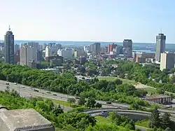 A tree-lined highway is in the foreground, angled diagonally from bottom right to middle left of the image. Buildings are in the centre, and the background is a sky meeting rolling hills in the distance.