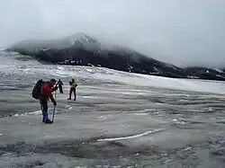 A black cone-shaped mountain rising over glacial ice and three climbers in the foreground