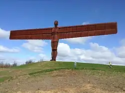 A large rust-coloured statue of a figure stretching wing-like arms out on top of a hill.