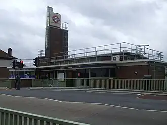 A red-bricked building with a blue sign reading "BOSTON MANOR STATION" and a person in the foreground all under a blue sky with white clouds