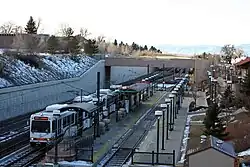 A white train is parked at a recessed island platformed station with canopies and lighting visible.