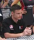 A man in his early twenties looking to someone outside the camera. He is wearing a black T-shirt with sponsors logos and his holding both his hands together/