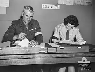 Two soldiers, one male, one female, sit side by side doing paper work at a desk. They are wearing brassards and the sign behind them says: "signal master".